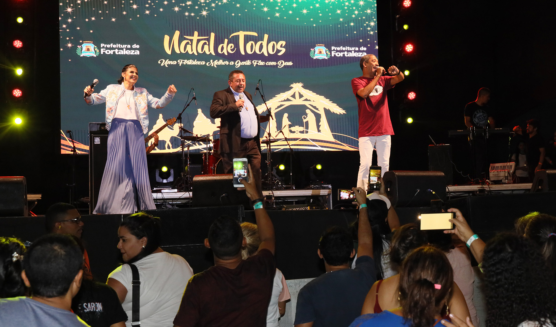 padre, mulher e homem cantando em cima de palco e público embaixo